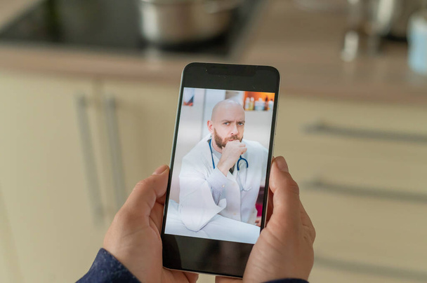 A serious bearded doctor communicates with a patient via a video call on a smartphone. Online medicine concept. Woman in the kitchen takes therapist remote consultation on mobile. - Fotó, kép