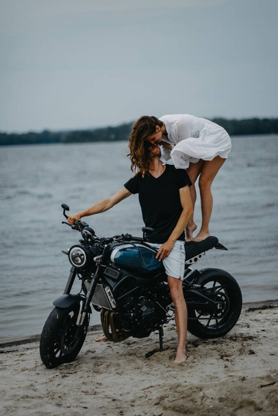 Liebespaar küsst sich am Strand in der Nähe von Motorrad auf Wasser Hintergrund. - Foto, Bild