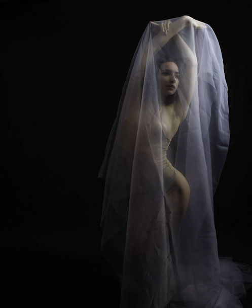 a girl of European appearance in a beautiful pose under a transparent fabric on a black background in a modern style - Foto, Imagen
