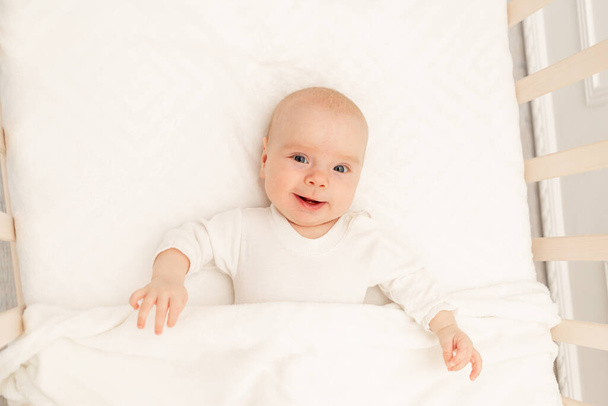 Το κοιμισμένο μωρό και το παιχνίδι του σε άσπρη κούνια. Βρεφοκομείο και κρεβάτια για παιδιά. Χαριτωμένο αγοράκι που κοιμάται στην κούνια. Το παιδί παίρνει έναν υπνάκο στο λευκό υπνοδωμάτιο. Υγιές παιδί με πιτζάμες.. - Φωτογραφία, εικόνα