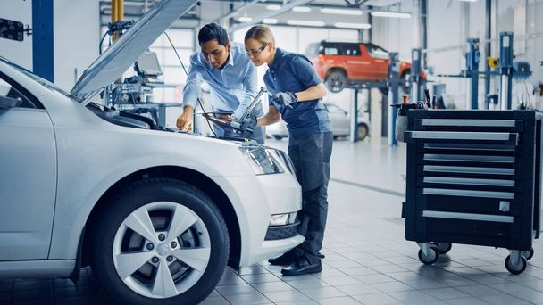 Manager Checks Diagnostics Results on a Tablet Computer and Explains an Engine Breakdown to an Female Mechanic. Car Service Employees Inspect Cars Engine Bay with a LED Lamp. Modern Clean Workshop. - Foto, Imagen