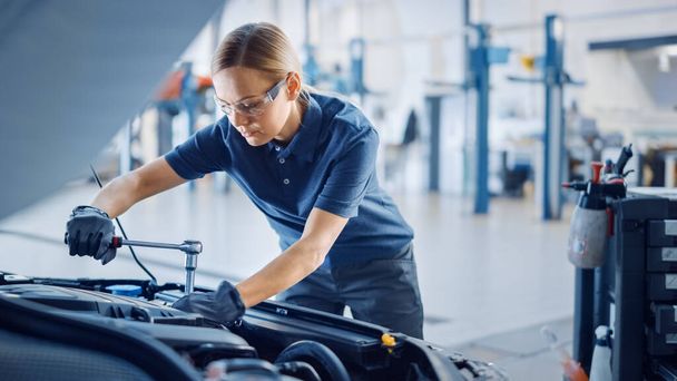 Beautiful Empowering Female Mechanic is Working on a Car in a Car Service. Woman in Safety Glasses is Working on an Usual Car Maintenance. Shes Using a Ratchet. Modern Clean Workshop with Cars. - Foto, imagen