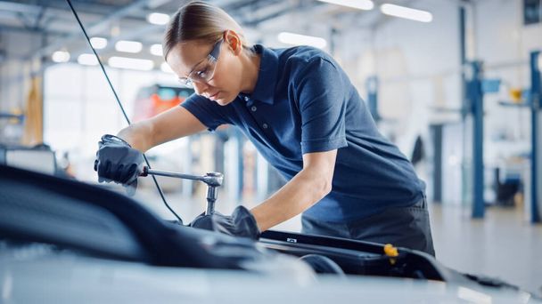 Beautiful Empowering Female Mechanic is Working on a Car in a Car Service. Woman in Safety Glasses is Working on an Usual Car Maintenance. Shes Using a Ratchet. Modern Clean Workshop with Cars. - Foto, Imagem