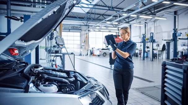 Female Mechanic Uses a Tablet Computer with an Augmented Reality Diagnostics Software. Specialist Inspecting the Car in Order to Find Broken Components Inside the Engine Bay. Modern Car Service. - Zdjęcie, obraz
