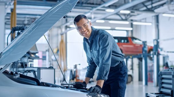 Handsome Professional Car Mechanic is Working on a Vehicle in a Service. Repairman Looks Happy While Using a Ratchet. Specialist is Wearing Safety Glasses. He Looks at a Camera and Smiles. - Photo, Image