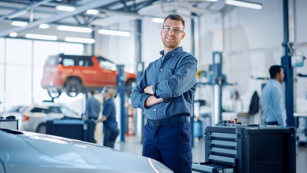Handsome Car Mechanic is Posing in a Car Service. He Wears a Jeans Shirt and Safety Glasses. His Arms are Crossed. Specialist Looks at a Camera and Smiles. - 写真・画像