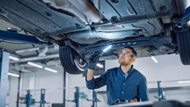 Handsome Professional Car Mechanic is Investigating Rust Under a Vehicle on a Lift in Service. Repairman is Using a LED lamp and Walks Towards. Specialist is Wearing Safety Glasses. Modern Workshop. - Photo, image