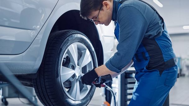 Mechanic in Blue Overalls is Unscrewing Lug Nuts with a Pneumatic Impact Wrench. Repairman Works in a Modern Clean Car Service. Specialists Removes the Wheel in Order to Fix a Component on a Vehicle. - Photo, Image