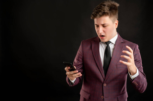 Law student with blond hair dressed in burgundy jacket, white shirt and black tie reading and sending text messages on cellphones displeased face posing on isolated black background with copy space advertising area - Photo, Image