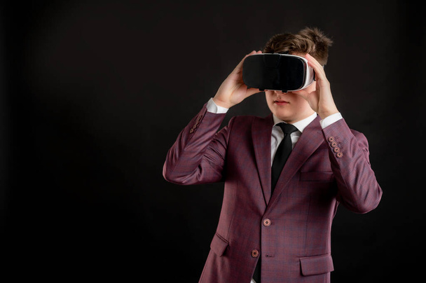 Estudiante de derecho con el pelo rubio vestido con chaqueta de color burdeos, camisa blanca y corbata negra con gafas de realidad virtual posando sobre un fondo negro aislado con área de publicidad de espacio de copia
 - Foto, Imagen