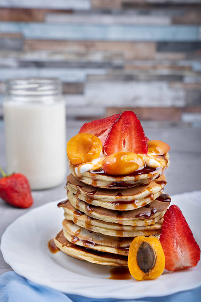 Pile of pancakes decorated with strawberries, apricots slices with syrup topping laying on white plate against background of jar with milk at kitchen - Фото, изображение