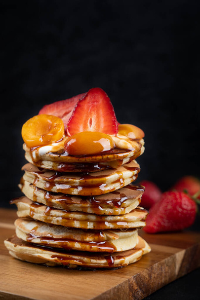 Stack of pancakes decorated with strawberry and apricot slices and with caramel syrup topping laying on wooden cutting board together with whole strawberries against dark background - Фото, изображение