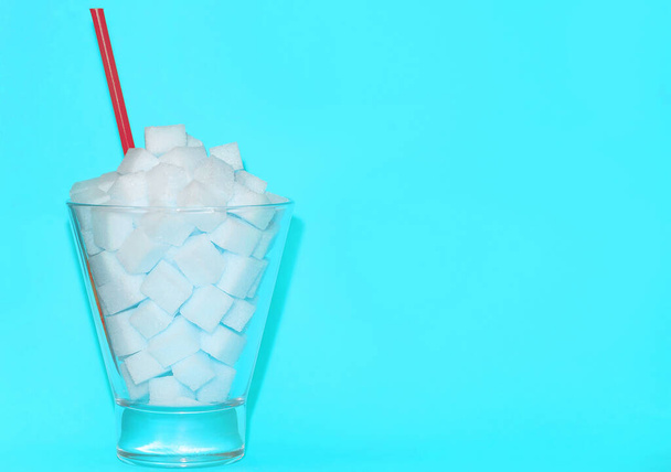 Glass with blue straw filled with sugar cubes on a pink background. Junk food concept, diabetes, a full glass of sweet water - Photo, image