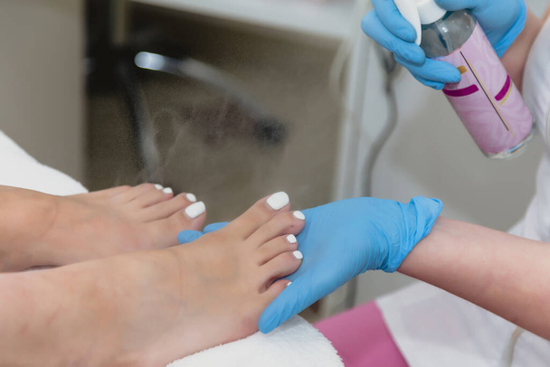 Pedicure master in blue rubber gloves sprays client feet before foot nail care procedure. Closeup. Pedicure in beauty salon. Hygiene and foot care. Concept of beauty industry - Fotó, kép