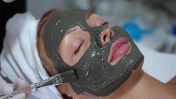 Il cosmetologo applica la maschera di argilla con pennello su faccia di donna carina giovane in clinica di bellezza, salone di centro benessere. Estetista fare bellezza facciale procedura di cura della pelle caucasica europa femminile
     - Filmati, video