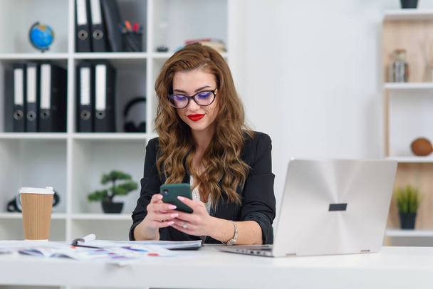 Smiling young business woman in stylish clothes sitting in cozy modern office and browsing something on smartphone. - Foto, imagen
