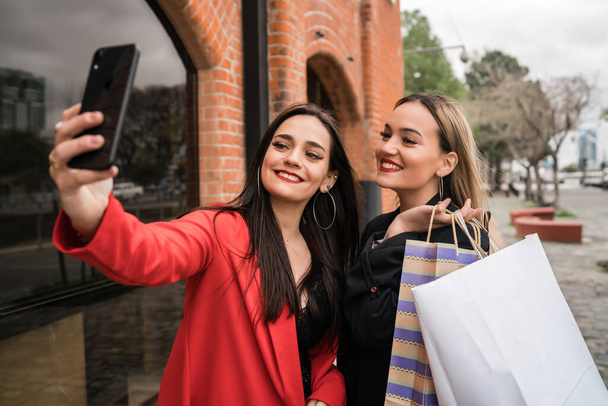 Portrait of two young friends enjoying doing shopping together while taking a selfie with phone at the street. Friendship and shopping concept. - Fotoğraf, Görsel