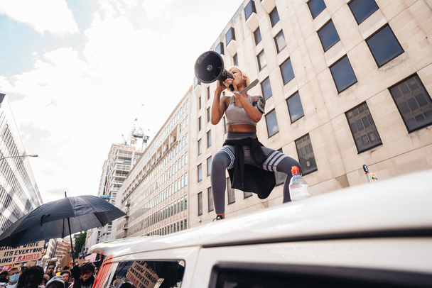Londyn / UK - 06 / 20 / 2020: Piękna dziewczyna z megafonem stojąca na dachu furgonetki przed tłumem protestujących w sprawie Black Lives Matters kierujących się na Parliament Square, Westminster, śpiewając i trzymając transparenty - Zdjęcie, obraz