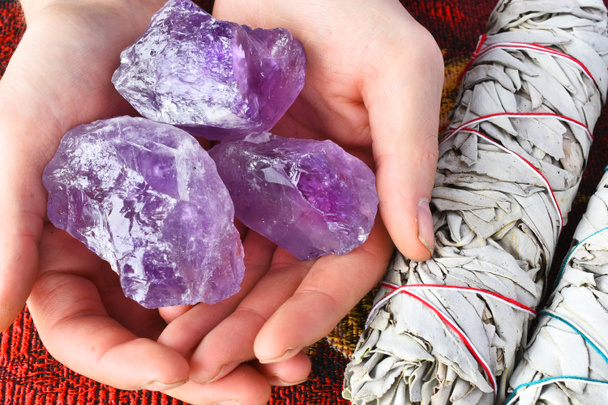 Nahaufnahme einer Hand, die drei lila Amethyst-Kristalle neben zwei weißen Salbeibündeln hält.  - Foto, Bild