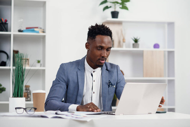 Serious dark-skinned young office manager works with different reports and table laptop at his workplace in the meeting room at modern business centre. - 写真・画像