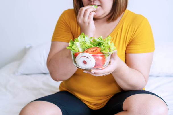 Az elhízott fiatal nők zöld organikus zöldségeket esznek, miközben a fehér ágyon ülnek. Kövér nő kezében tál saláta vegetáriánus, miközben eszik saláta a kezében. Koncepció élelmiszer jó egészséges fogyás. - Fotó, kép
