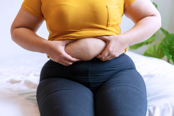 Overweight young woman pinches fat on her belly large size while sitting on white bed at home. Upset female suffering from extra weight. Obesity unhealthily concept. - Φωτογραφία, εικόνα