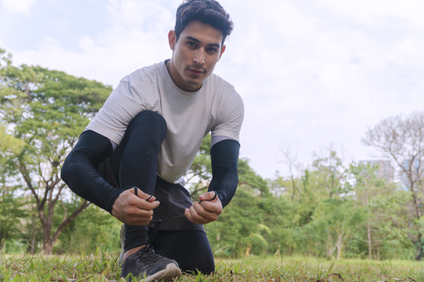 Athletic young man kneeling while tying sports shoes in the park looking at camera. Handsome male shoelace on green grass with big tree in the park. Healthy workout concept. - Photo, Image
