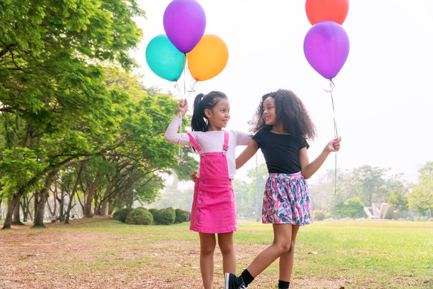 Wesoła dwójka dzieci trzyma kolorowe balony stojąc razem na zielonej trawie. Cute lovely girl grać z balonami uczucie wolności, patrząc na siebie w parku. Koncepcja przyjaźni. - Zdjęcie, obraz