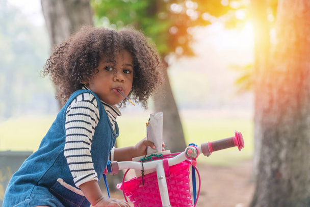 Little 2.6 years old black girl want learn ride bicycle eating chocolate in her mouth while standing in the park with tree. Mixed race kid girl hold pink small bike  at outdoor on summer day.  - Foto, afbeelding