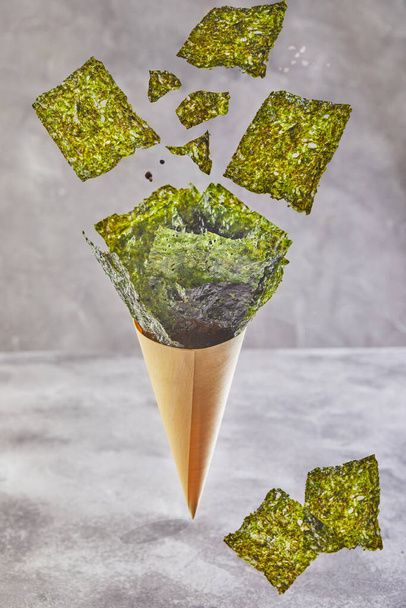 Crispy nori seaweed falling in a cone cup on a gray background, levitation. Japanese food nori. Dried sheets of seaweed. - Photo, Image