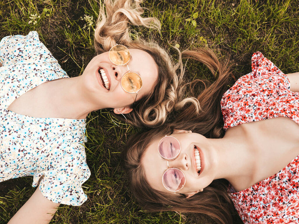 Two young beautiful smiling hipster girls in trendy summer sundress.Sexy carefree women lying on the green grass in sunglasses.Positive models having fun.Top view - Foto, Bild