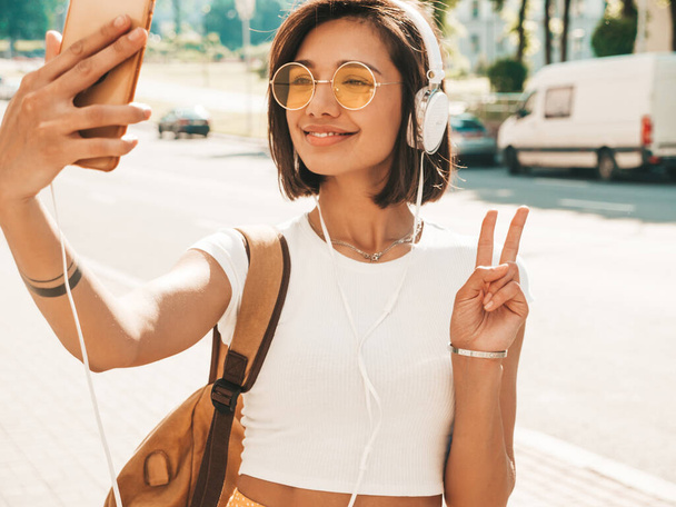 Fashion portrait of young stylish hipster woman walking in the street.Girl making selfie and shows peace sign.Smiling model enjoy her weekends with backpack. Female listening to music via headphones - 写真・画像