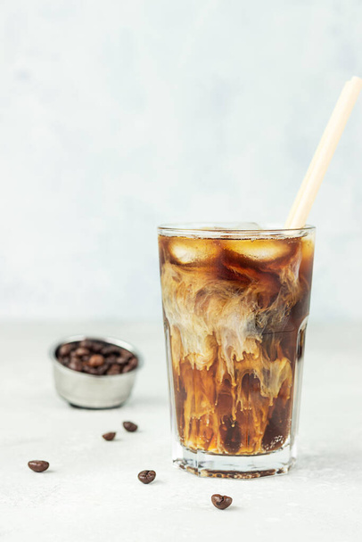 Iced coffee with milk in a tall glass with roasted coffee beans on light grey background. Summer refreshment drink. Selective focus. - Φωτογραφία, εικόνα