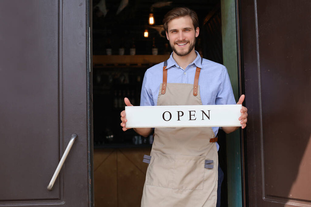 Smiling male owner of coffee shop is standing outdoors. He is holding placard and inviting everybody to come in. Open - Фото, изображение