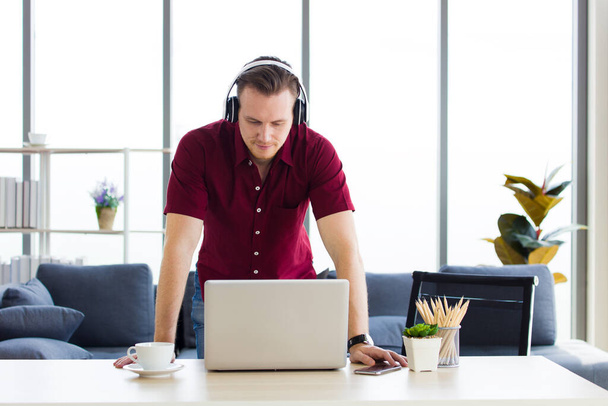 Attractive man wearing earphones is chatting with colleagues via video call on the laptop at home during having outbreaks of coronavirus Covid-19. - 写真・画像