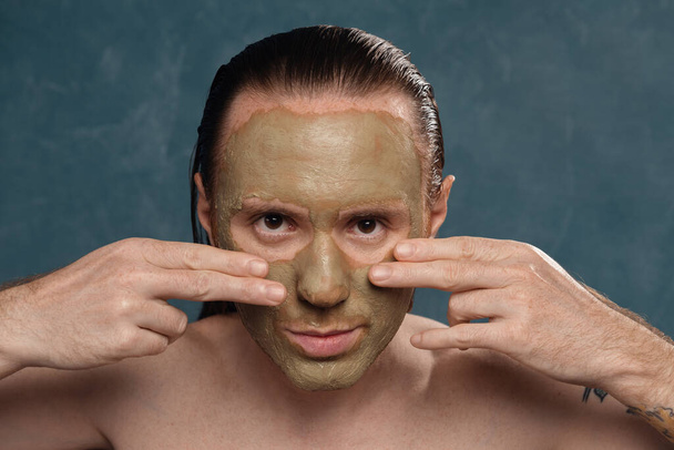 Татуированный мужчина с длинными мокрыми волосами надевает зеленую глиняную маску руками
. - Фото, изображение