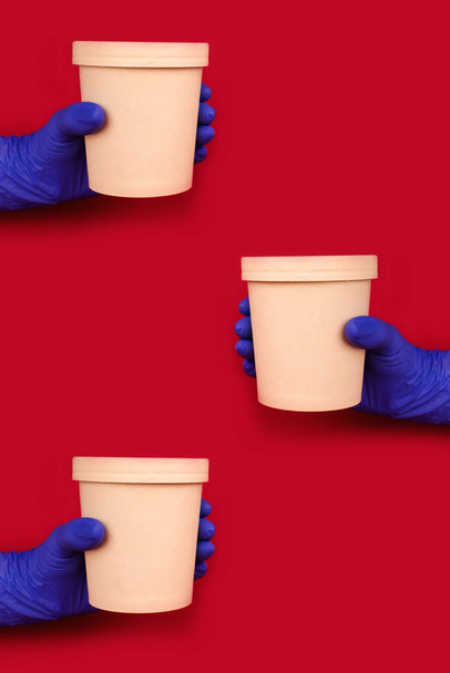 Κάθετη σύνθεση με τα χέρια να φορούν μπλε γάντια μιας χρήσης σε κόκκινο φόντο κρατώντας στρογγυλό χάρτινο δοχείο τροφίμων - χάρτινο κύπελλο για σούπα ή άλλα πιάτα takeaway. Copyspace, μακιγιάρισμα - Φωτογραφία, εικόνα