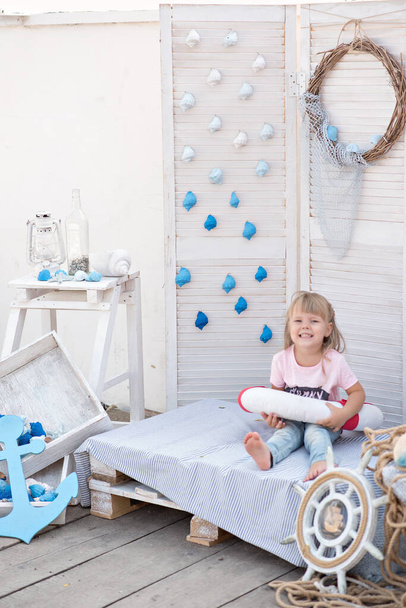 Beautiful little blonde girl with blue eyes looks at the camera and laughs. The baby plays with a lifebuoy and shells on the background of marine decoration. Sea adventures and games for children.  - Φωτογραφία, εικόνα