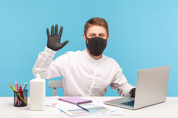 Friendly male employee working on laptop and gesturing hello to camera, wearing mask, hygienic gloves to prevent coronavirus at workplace, protection against contagious disease in office. studio shot - Photo, image