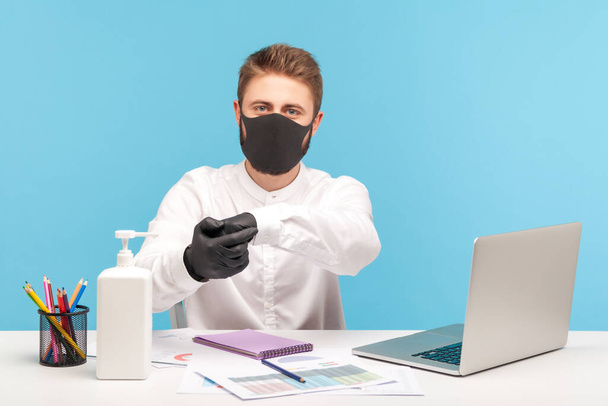 Male employee in hygienic mask and gloves rubbing hands, using antiseptic gel, disinfection to prevent coronavirus at workplace, protection against contagious disease in office. studio shot isolated - Photo, image