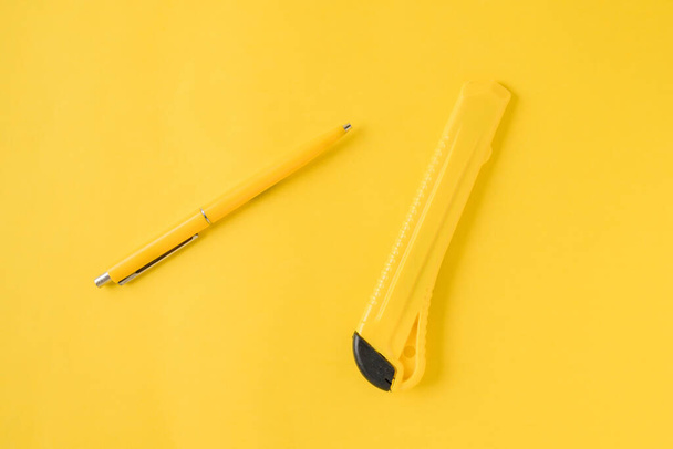 Yellow pen and office knife on the yellow background. Top view. Minimalism. Clean looking background. Flat lay. Template with copy space. Two objects. Concept chancellery, office supplies, accessories - Foto, Imagem