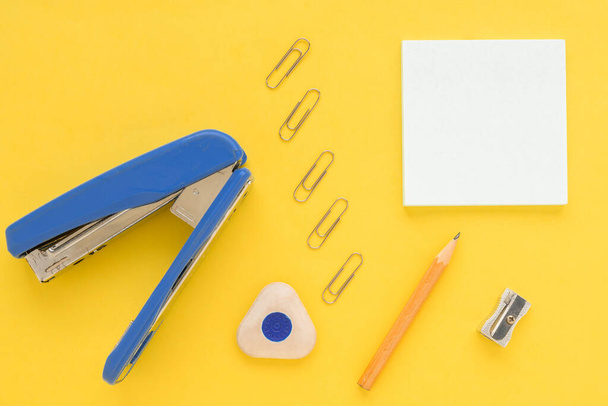 White square notes, pencil, eraser, blue stapler, sharpener, metal paper clip on yellow paper background - Foto, afbeelding