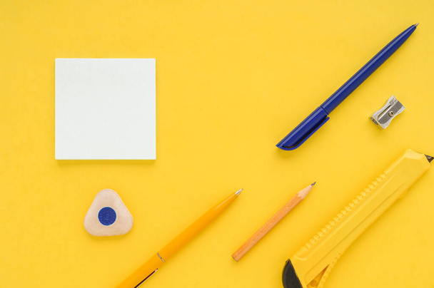 White square notes, pencil, eraser, sharpener, office knife, blue and yellow pen on yellow paper background - Фото, изображение