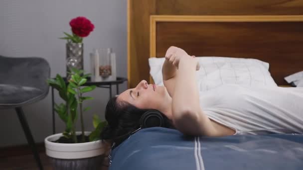 Beautiful relaxing satisfied woman lying on bed and enjoying modern music in headphones,slow motion - Felvétel, videó