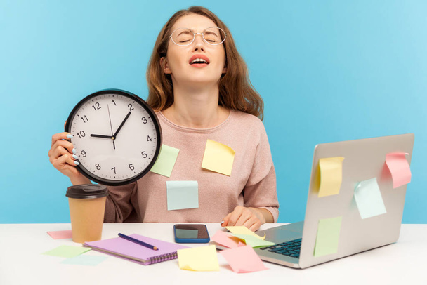 Overtime work. Fatigued upset woman employee sitting at workplace office, all covered with sticky notes and holding big clock, screaming from despair and exhaustion. indoor studio shot, isolated - Photo, image