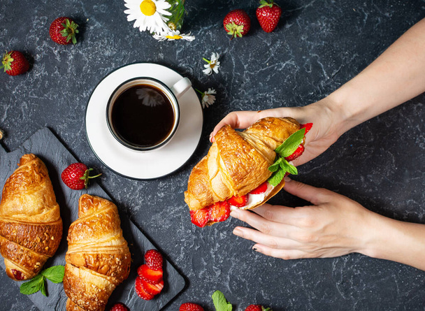 Tasty breakfast. Female hands holds fresh croissants with strawberries on black stone background. Top view - Photo, Image