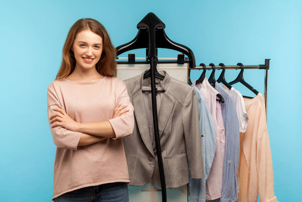 Confident happy woman, professional expert fashion stylist standing near clothes hang on shelf in designer store, selling trendy outfit in shop boutique. indoor studio shot isolated on blue background - Φωτογραφία, εικόνα