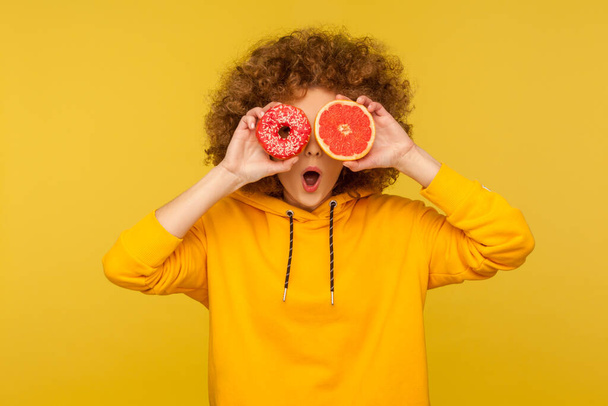 Fruits vs sugary desserts. Portrait of surprised curly-haired woman in urban style hoodie covering eyes with fresh grapefruit and sweet doughnut, open mouth in amazement. indoor studio shot isolated - Foto, Imagem