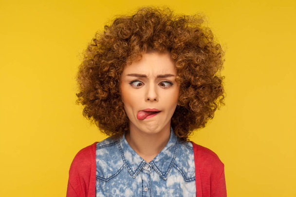 Closeup portrait of crazy woman with fluffy curly hair demonstrating tongue out and making silly face with crossed eyes, dumb brainless expression. indoor studio shot isolated on yellow background - Photo, Image