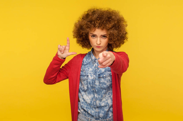 Portrait of curly-haired woman expressing disrespect, showing L finger sign, loser or lame gesture and pointing to camera, accusing for failure. indoor studio shot isolated on yellow background - Photo, Image